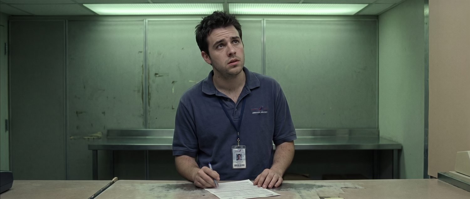 Medium shot of Trevor, mid-twenties in a navy airline uniform polo, standing behind the counter with a printed inventory form squared in front of him and a pen beside it, lit from directly overhead.