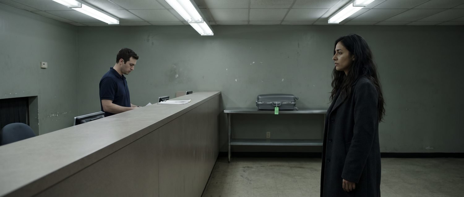 Alternate wide shot of the same baggage service counter, with a different actress in the Elise role. Same institutional geometry, same suitcase on the shelf behind Trevor.