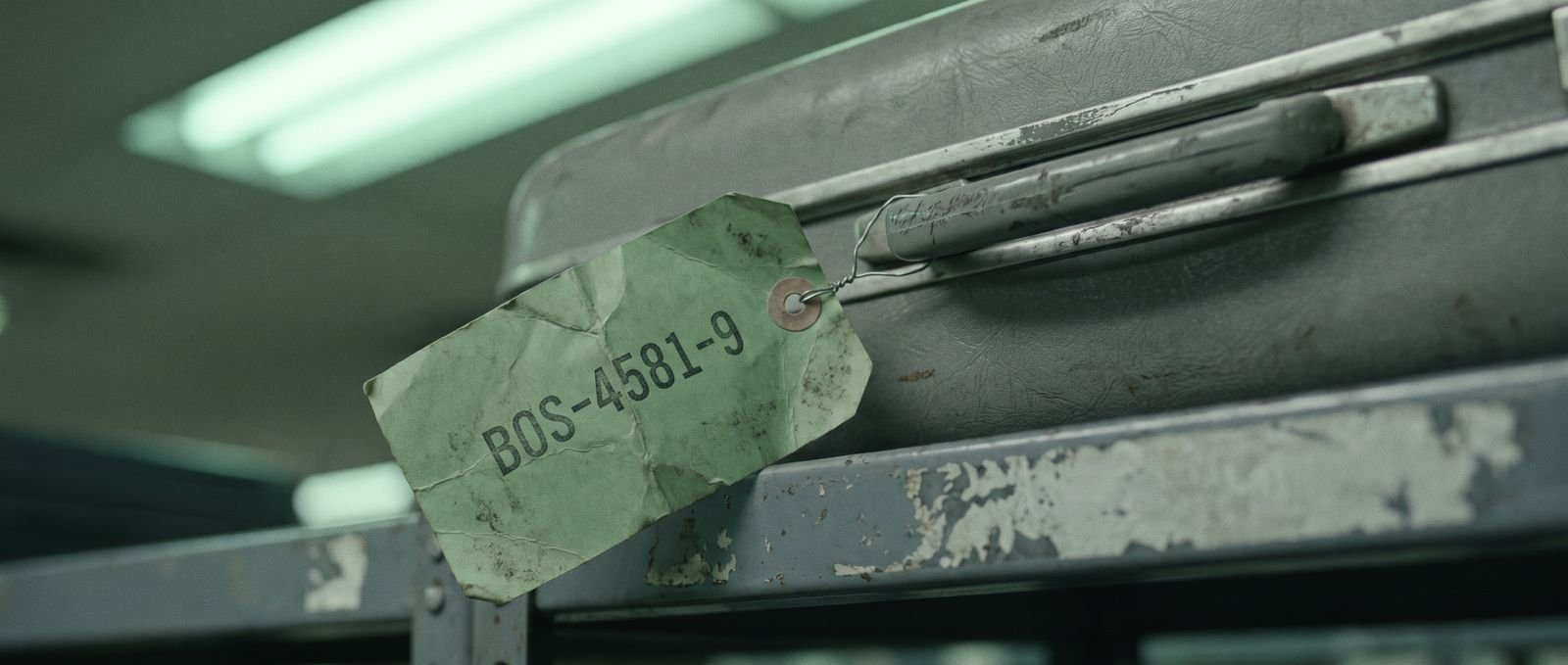 A green paper baggage tag reading BOS-4581-9, wired to the handle of a grey hardshell suitcase on a steel shelf, under fluorescent light.
